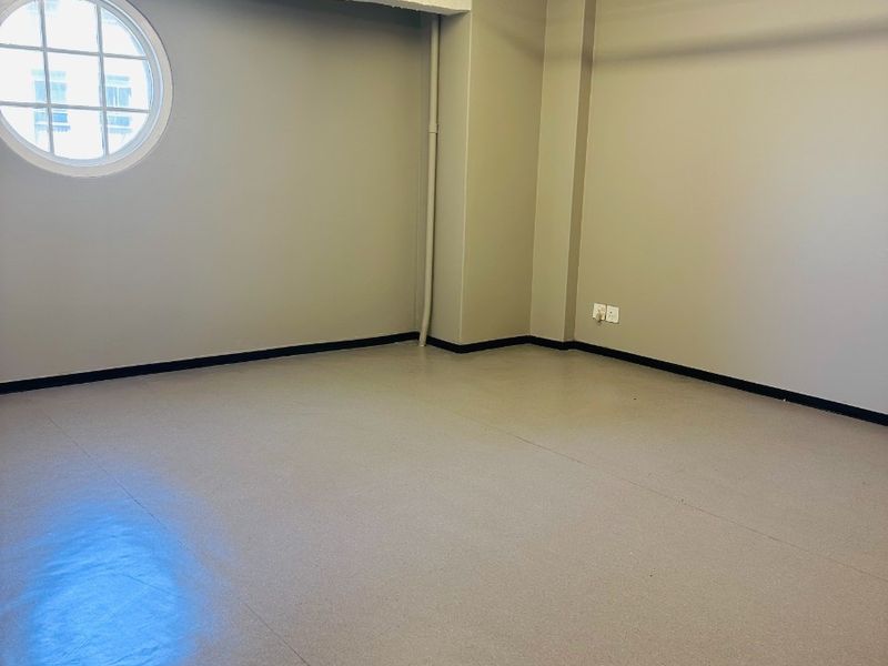 A clean, empty office space featuring industrial-style exposed ceiling beams, a distinctive circular window, and durable vinyl flooring with integrated fire safety infrastructure.
