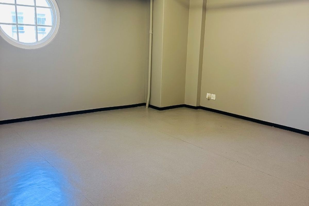 A clean, empty office space featuring industrial-style exposed ceiling beams, a distinctive circular window, and durable vinyl flooring with integrated fire safety infrastructure.