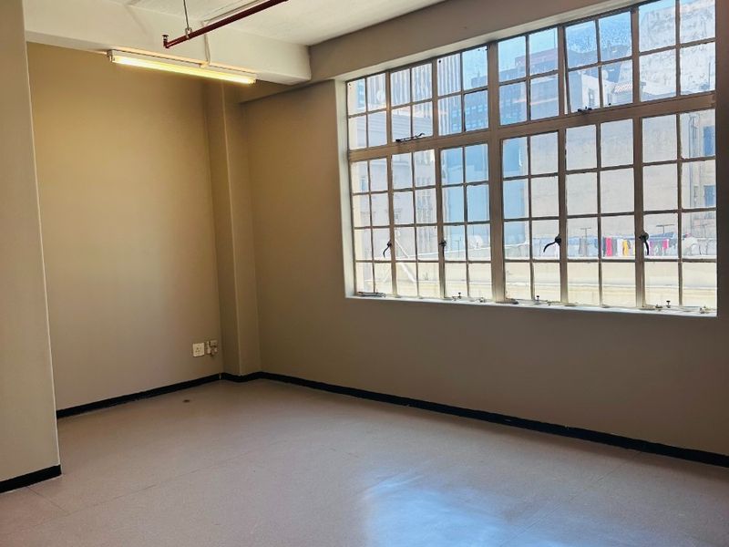 An empty, industrial-style office space featuring exposed concrete ceilings, large steel-framed windows, and functional strip lighting, suitable for a modern workspace.