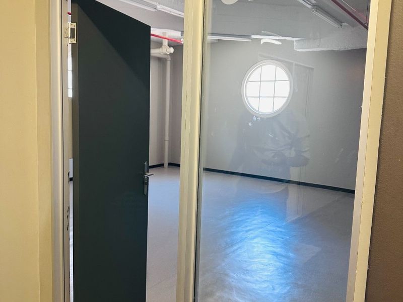 A vacant office space featuring a glass partition entrance, exposed ceiling infrastructure, and a distinctive circular window, finished with clean epoxy flooring.