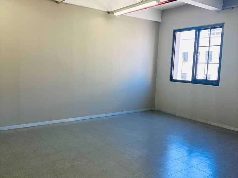 A bright, empty office space featuring exposed ceiling infrastructure, fire sprinkler system, tiled flooring, and a large window providing natural light.