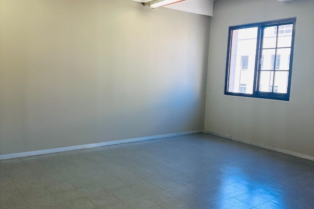A bright, empty office space featuring exposed ceiling infrastructure, fire sprinkler system, tiled flooring, and a large window providing natural light.