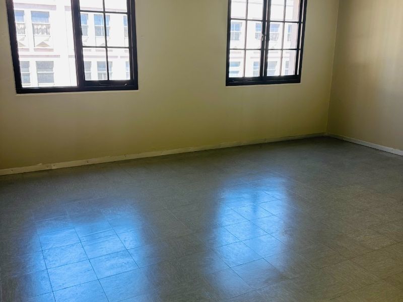 An empty office space featuring vinyl tile flooring, exposed fire sprinkler piping, and large grid windows providing natural light.