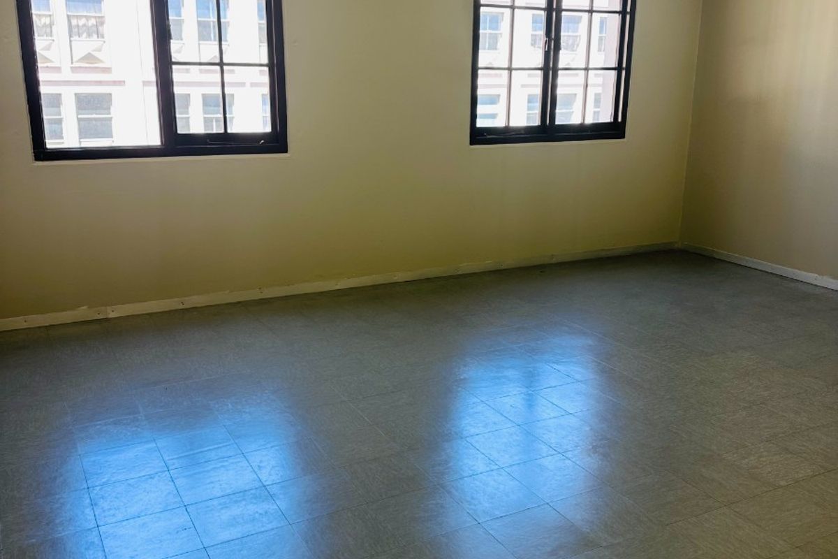 An empty office space featuring vinyl tile flooring, exposed fire sprinkler piping, and large grid windows providing natural light.