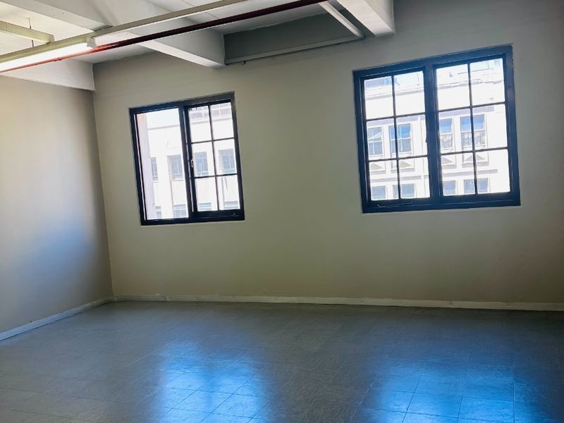 An empty, open-plan office space featuring exposed ceiling beams, fluorescent lighting, and large grid windows providing natural light.