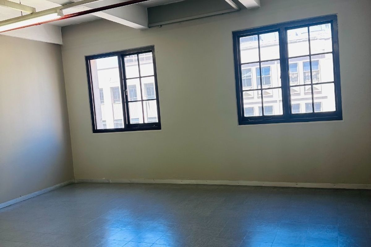 An empty, open-plan office space featuring exposed ceiling beams, fluorescent lighting, and large grid windows providing natural light.