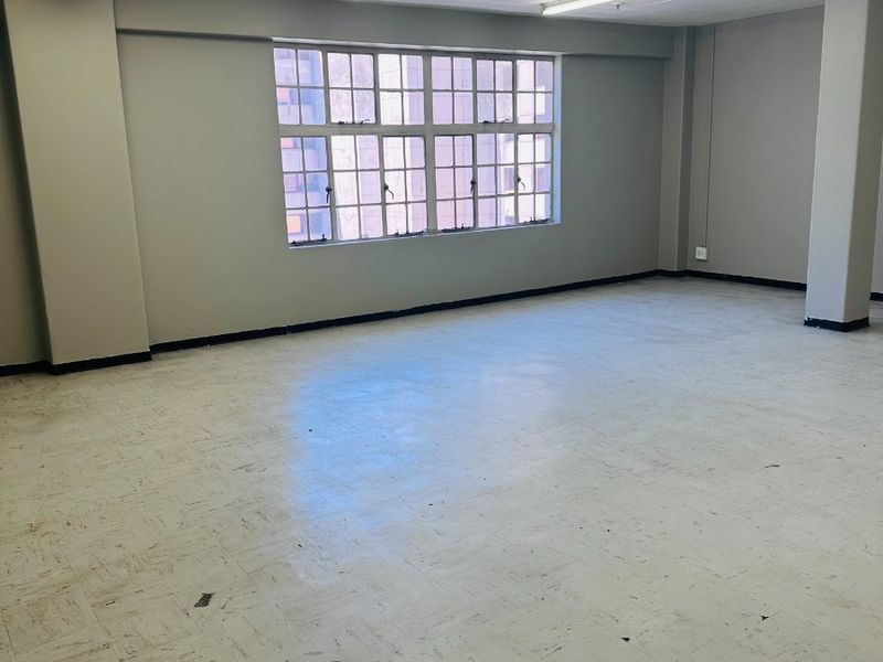 An empty, open-plan office space featuring vinyl flooring, grid ceiling with fluorescent lighting, and large steel-framed windows providing natural light.