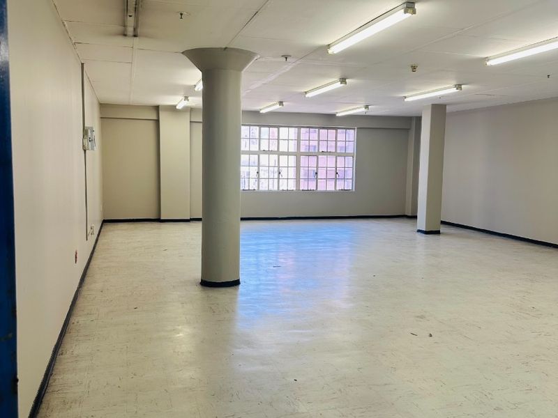 A spacious, empty open-plan commercial office area featuring structural columns, vinyl flooring, and ample natural light from multi-pane windows.
