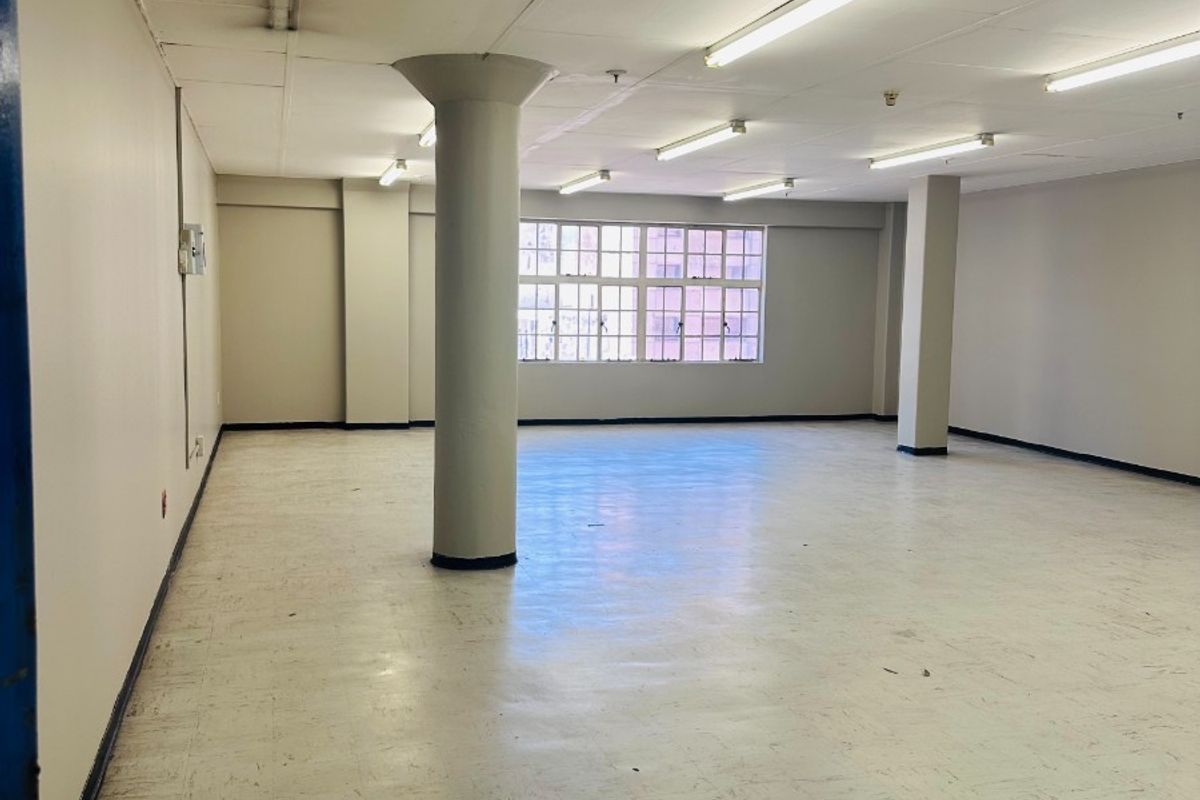 A spacious, empty open-plan commercial office area featuring structural columns, vinyl flooring, and ample natural light from multi-pane windows.