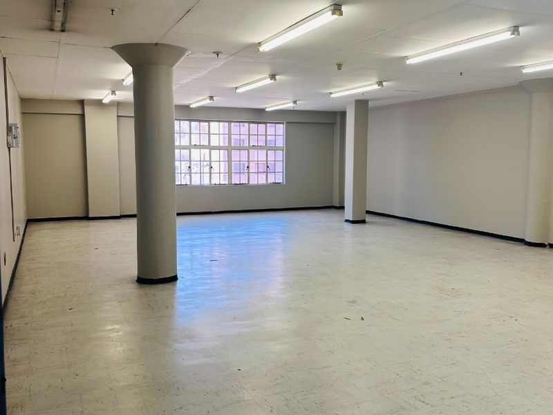 A spacious, empty open-plan commercial office area featuring structural columns, vinyl flooring, and fluorescent lighting, suitable for various business configurations.