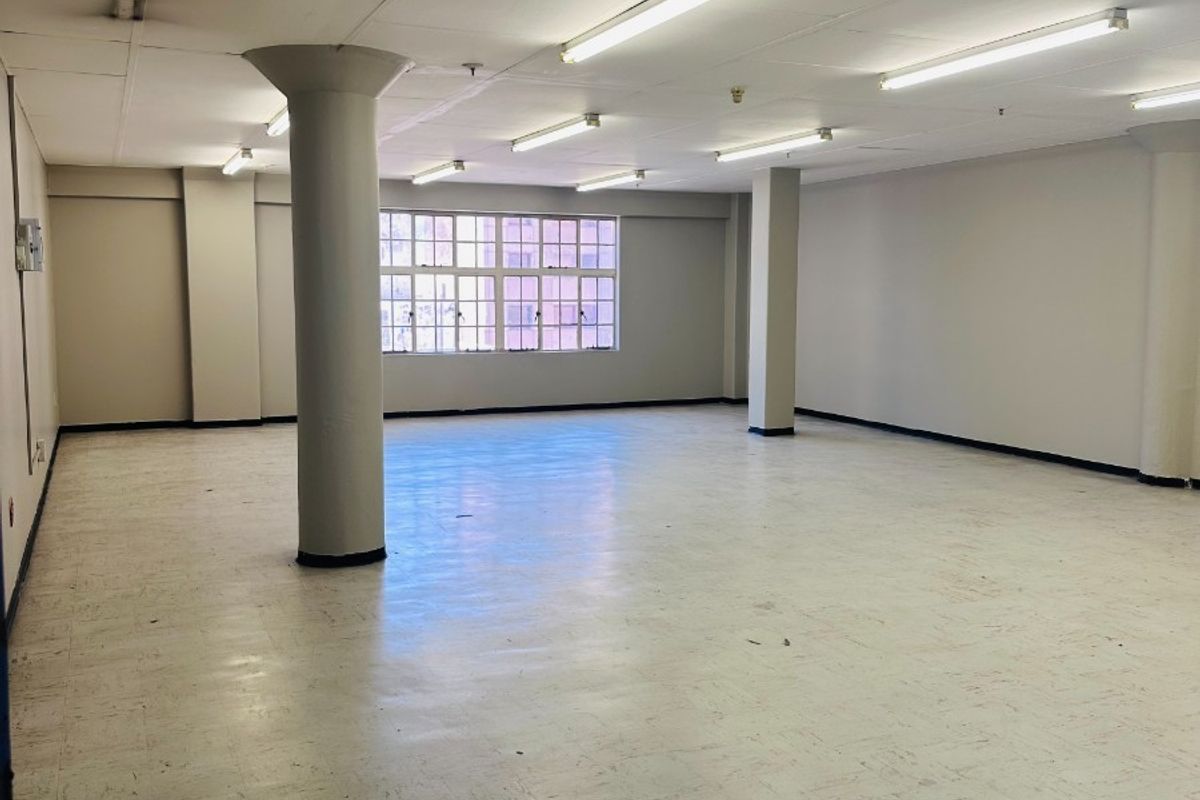 A spacious, empty open-plan commercial office area featuring structural columns, vinyl flooring, and fluorescent lighting, suitable for various business configurations.