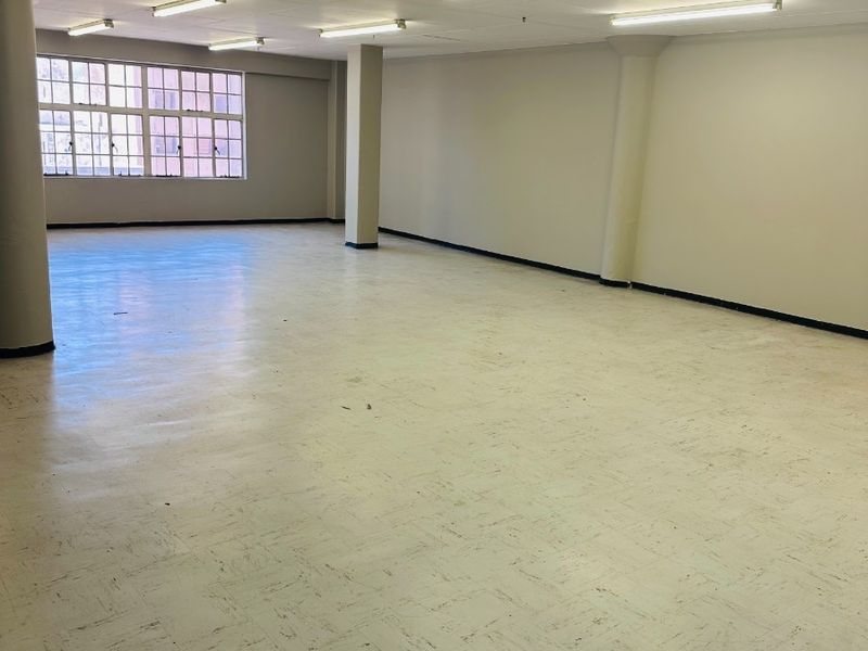 A spacious, empty open-plan office area featuring structural columns, fluorescent lighting, and large windows providing natural light.