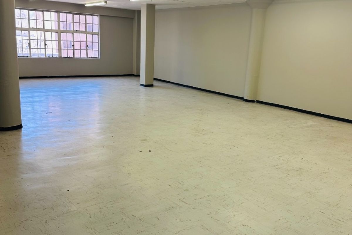 A spacious, empty open-plan office area featuring structural columns, fluorescent lighting, and large windows providing natural light.