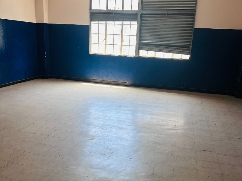 An empty office space featuring exposed ceiling pipes, grid windows with a damaged security shutter, and worn vinyl tile flooring.