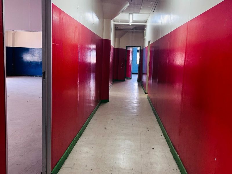 A long, vacant commercial hallway featuring vinyl tile flooring, exposed ceiling conduit, and high-contrast painted walls.