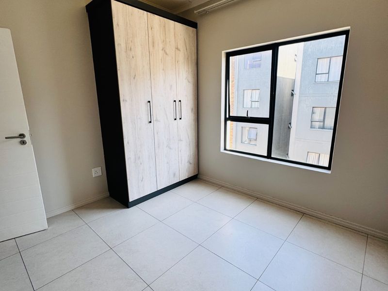 A bright, empty bedroom featuring neutral tiled flooring, a modern built-in wardrobe with wood-grain finish, and large aluminium-framed windows.