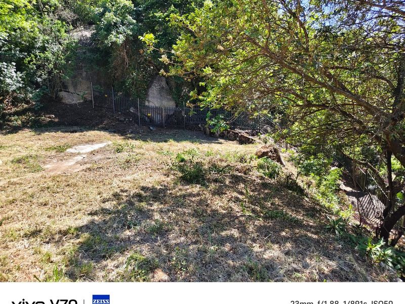 A sloped residential plot featuring natural rock formations, mature trees, and existing metal perimeter fencing.