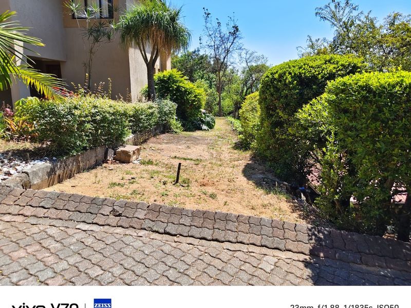 A residential garden area featuring a paved driveway edge, established hedges, and a sloped lawn area adjacent to the building structure.