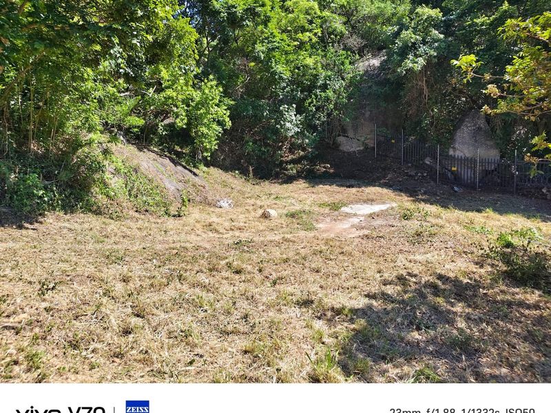 A vacant residential plot featuring natural rock formations, mature trees, and existing perimeter fencing.