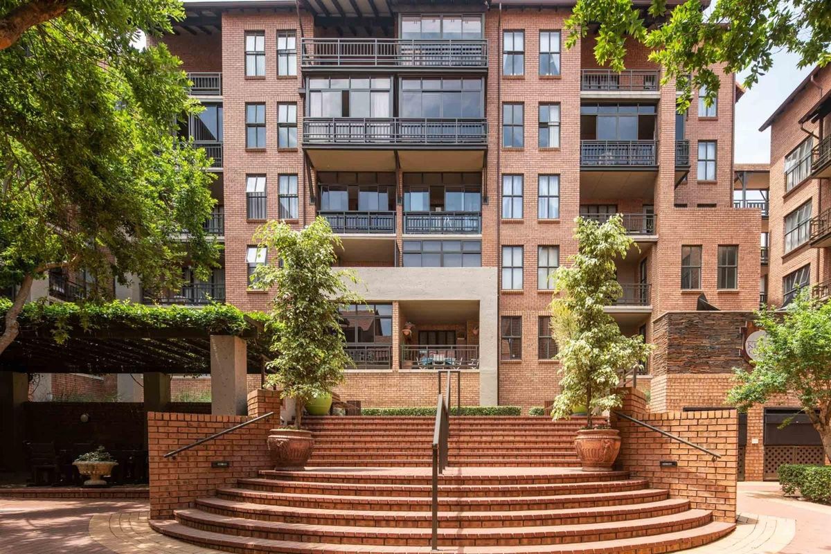A well-maintained multi-story apartment building featuring a prominent brick facade, tiered brick staircase, and private balconies with metal railings.