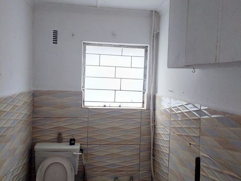 A compact bathroom featuring a bathtub, toilet, and wall-mounted storage cabinet, finished with patterned wall tiles and security bars on the window.