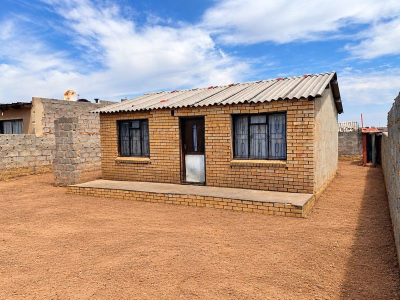 A detached single-story face brick house featuring a corrugated roof, security bars on windows, and a concrete porch, set on an open dirt plot.