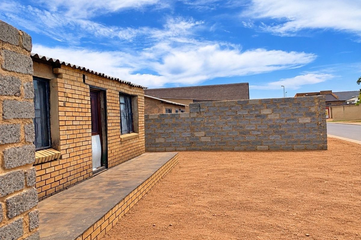 Exterior view of a face brick house featuring a paved walkway and an unfinished concrete block boundary wall on a large, undeveloped plot.