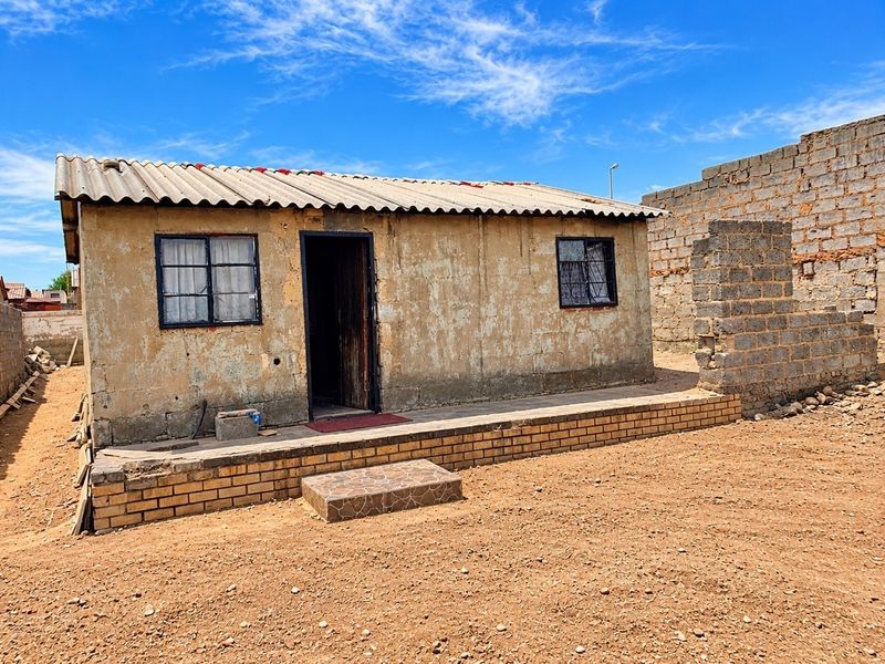 A detached single-story dwelling featuring a corrugated roof, brick foundation, and security-barred windows, set on an unpaved plot with an incomplete boundary wall.