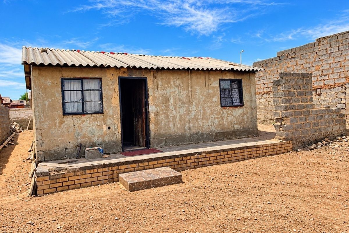 A detached single-story dwelling featuring a corrugated roof, brick foundation, and security-barred windows, set on an unpaved plot with an incomplete boundary wall.