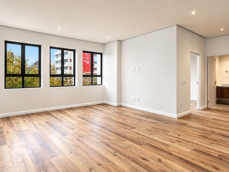 A bright, modern, and empty living area featuring high-quality laminate wood flooring, recessed downlights, and large black-framed windows with views.