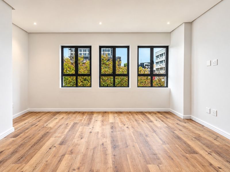 A bright, empty living room featuring modern laminate wood flooring, recessed downlights, and large black-framed windows with a pleasant urban view.