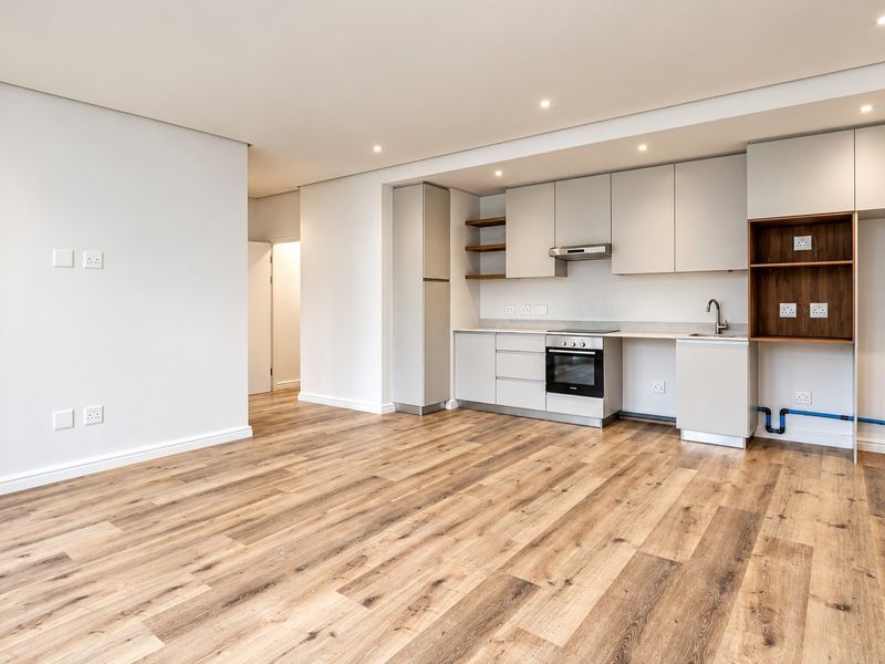 Modern open-plan living area featuring light-toned laminate flooring, recessed lighting, and a compact kitchen with built-in cabinetry, oven, and extractor fan.