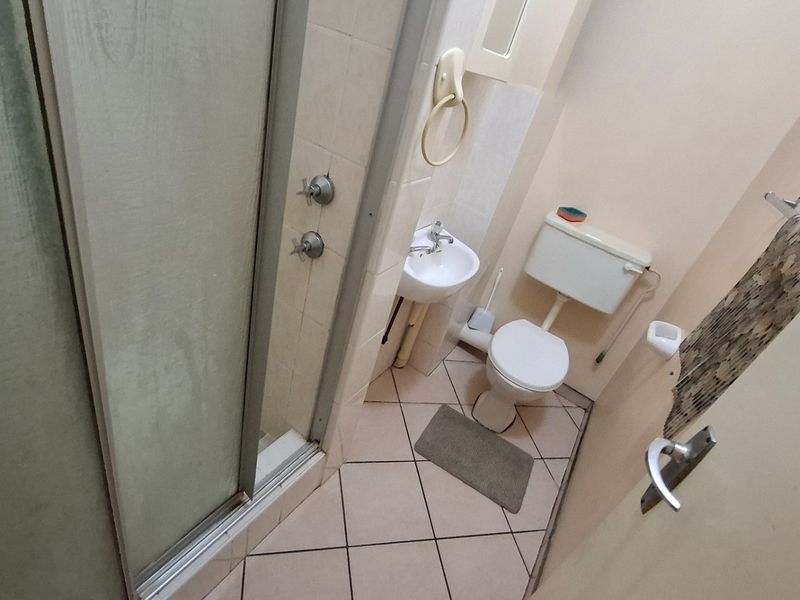 A compact bathroom featuring a shower enclosure, wall-mounted basin, and toilet with dated ceramic floor tiling and neutral wall finishes.