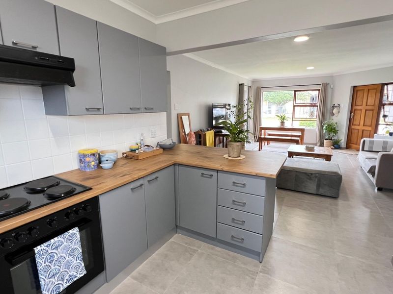 Modern open-plan kitchen featuring grey cabinetry, wooden countertops, and integrated appliances, flowing seamlessly into a bright and spacious living area with tiled floors.