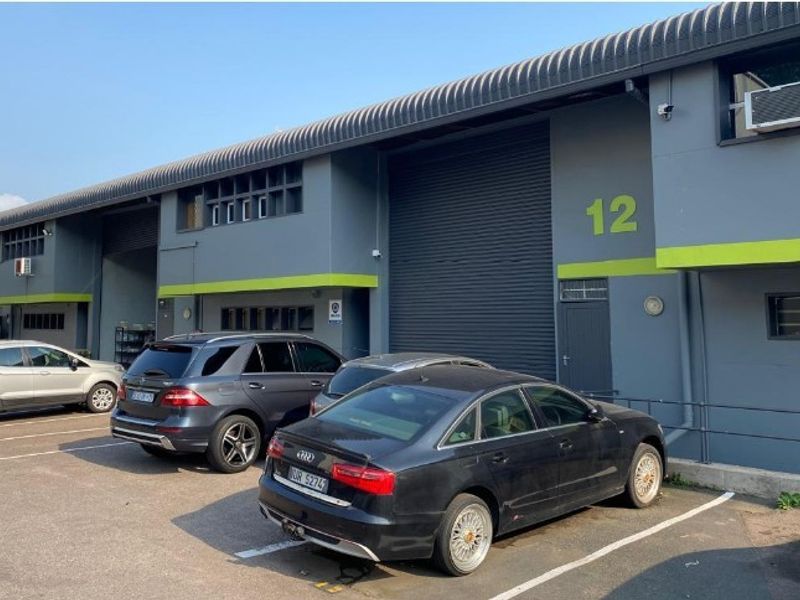 Front elevation of an industrial unit featuring a roller shutter door, grey facade with green accents, and dedicated asphalt parking space.