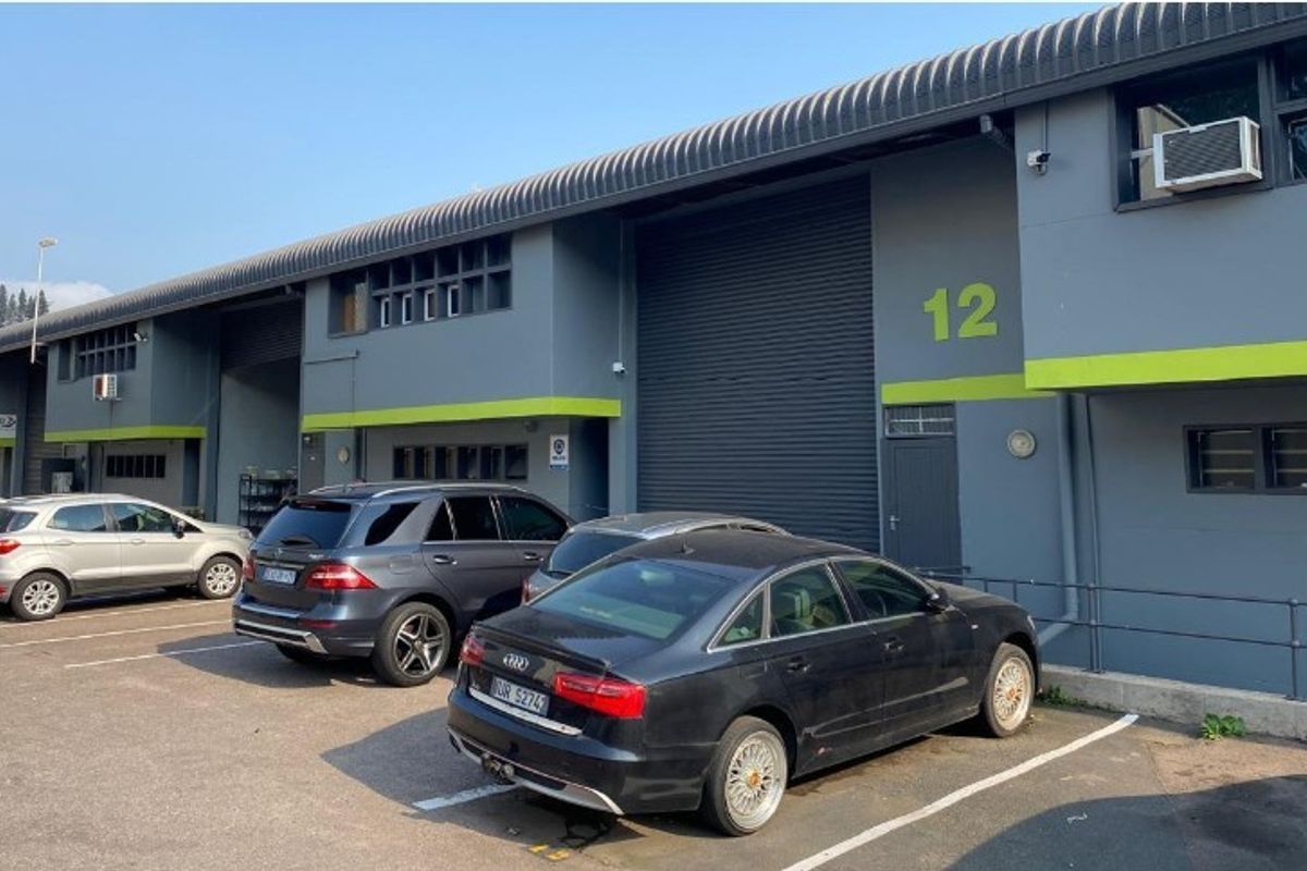 Front elevation of an industrial unit featuring a roller shutter door, grey facade with green accents, and dedicated asphalt parking space.