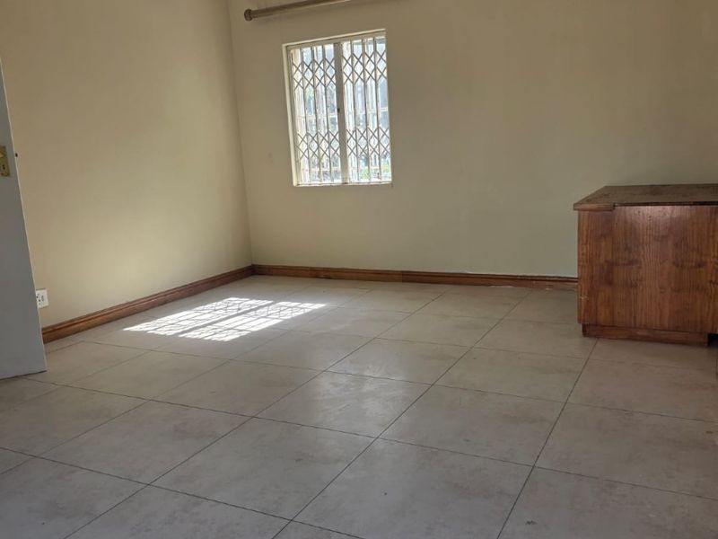 A spacious, empty living area featuring neutral tiled flooring, recessed lighting, and a window fitted with a security gate.