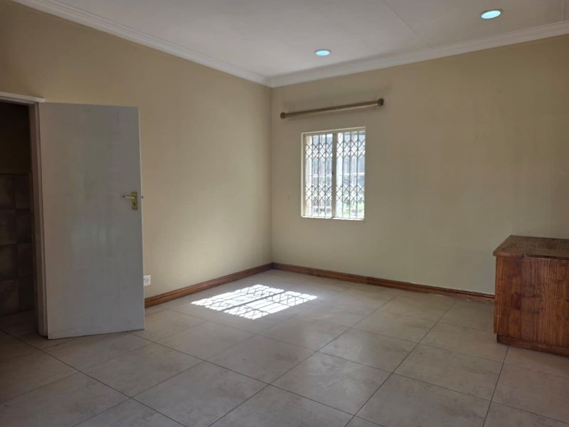 A clean, empty bedroom featuring neutral wall tones, tiled flooring, recessed lighting, and a window fitted with a security gate.