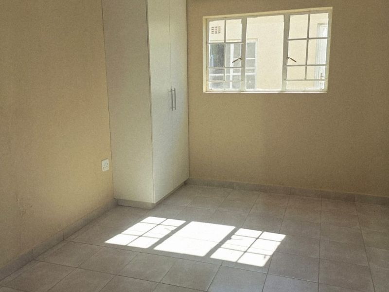 A bright, empty bedroom featuring neutral tiled flooring, a built-in wardrobe, and recessed ceiling lighting, offering a clean canvas for personalization.