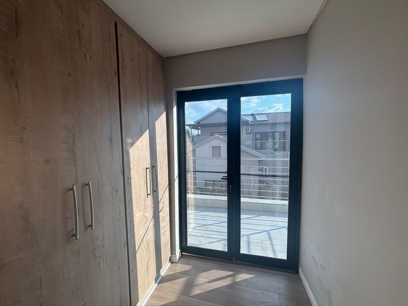A bright, empty bedroom featuring modern wood-finish built-in cupboards, laminate flooring, and double glass doors leading out to a balcony.