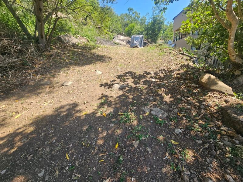 A sloped, undeveloped residential plot featuring natural rock formations and mature trees, offering potential for landscaping or future construction.