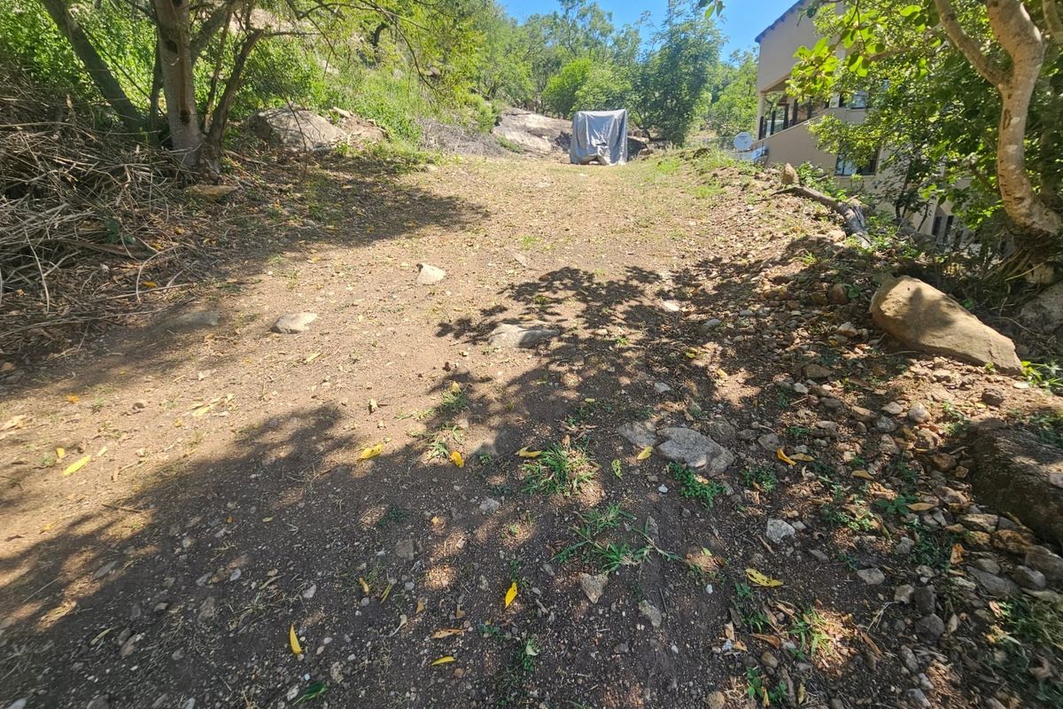 A sloped, undeveloped residential plot featuring natural rock formations and mature trees, offering potential for landscaping or future construction.