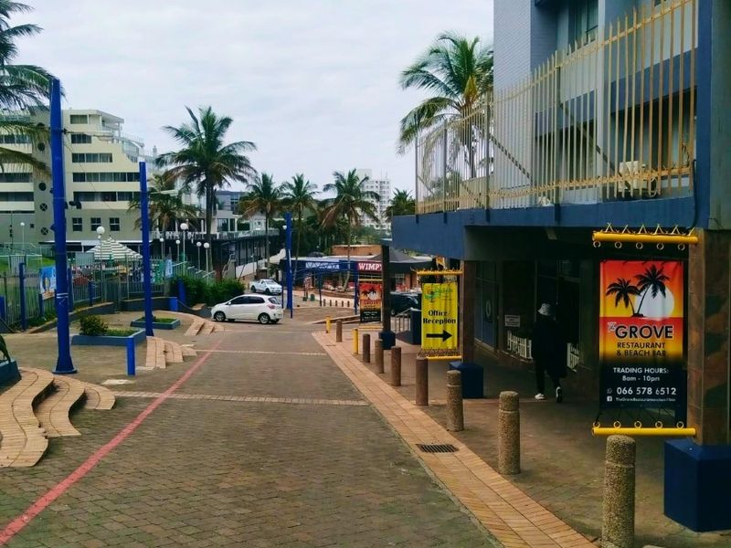 A street-level view of a commercial property entrance featuring paved walkways, bollards, and prominent signage for The Grove Restaurant and Beach Bar.