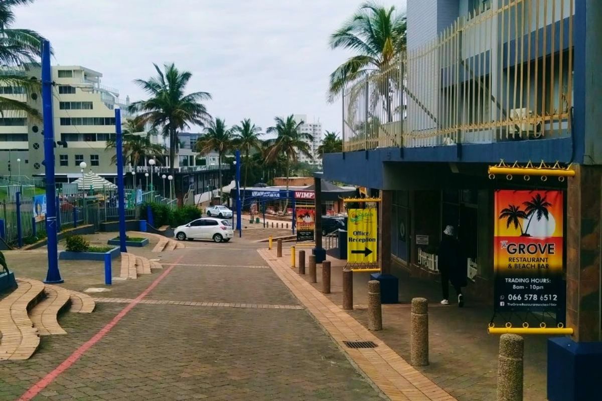 A street-level view of a commercial property entrance featuring paved walkways, bollards, and prominent signage for The Grove Restaurant and Beach Bar.