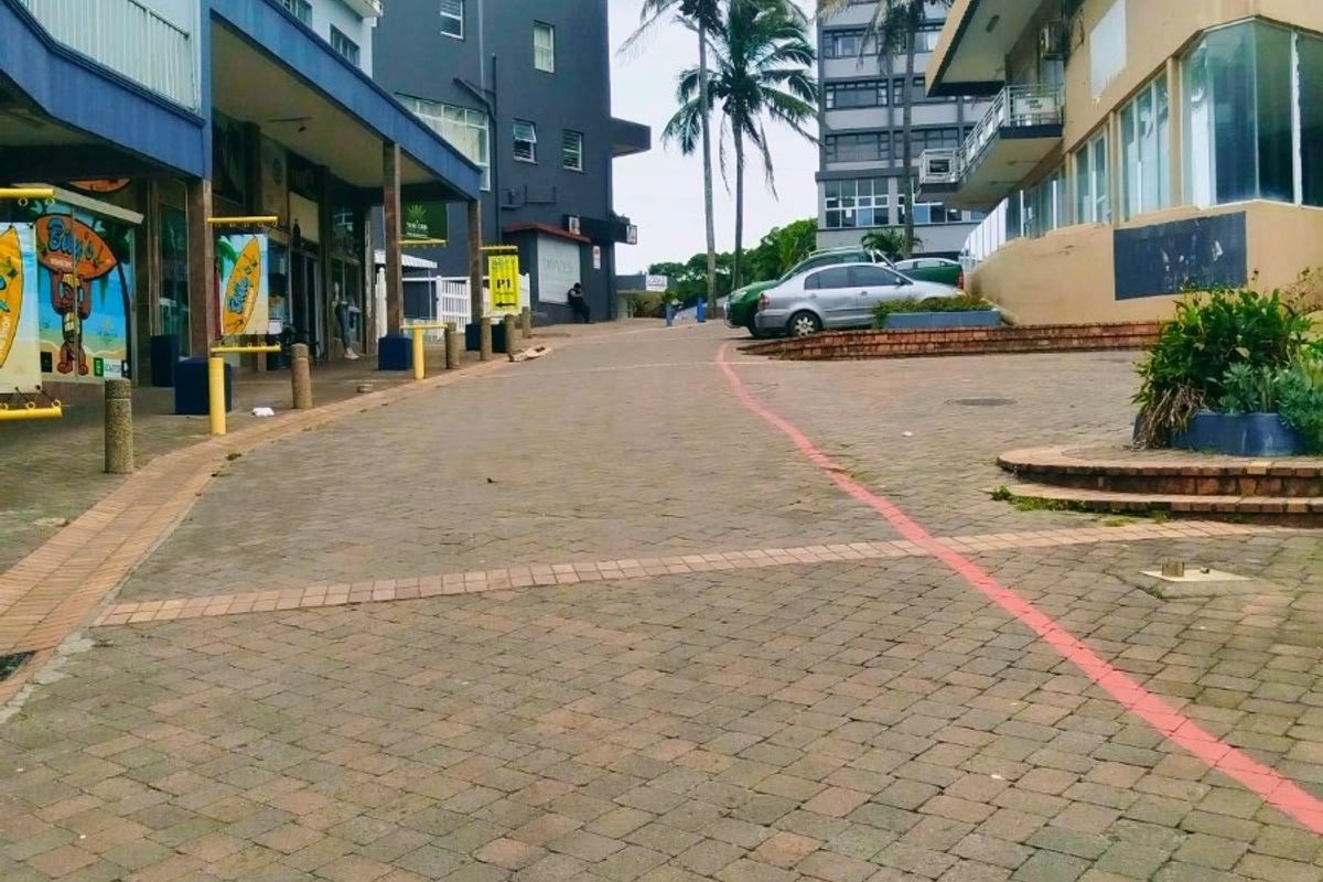 A street-level view of a commercial area featuring paved walkways, multi-story buildings, and storefronts with visible signs of exterior wear.