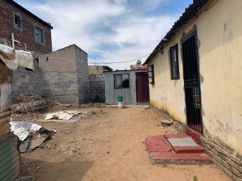 Rear exterior view of the property showing a dirt yard, basic brick and corrugated metal structures, and a security gate entrance.