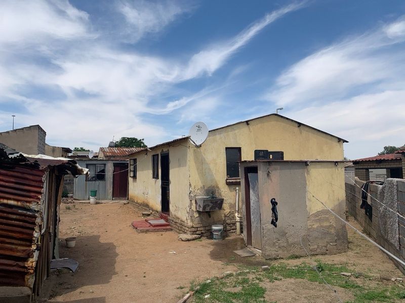 Rear view of a residential property showing exterior walls with visible plaster damage and basic utility fixtures in a yard setting.