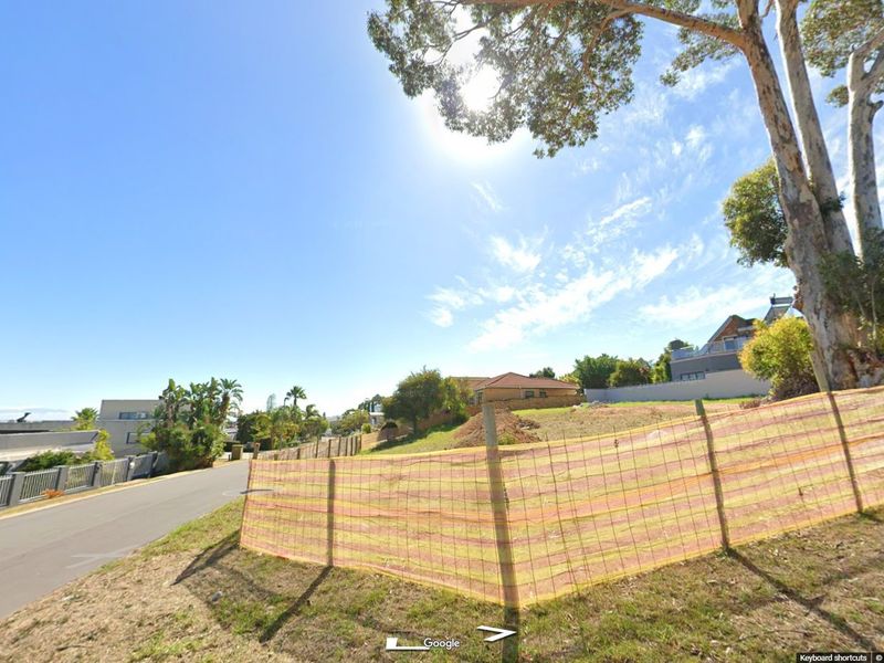 A street-level view of a vacant residential plot featuring perimeter fencing and established trees in a suburban neighborhood.