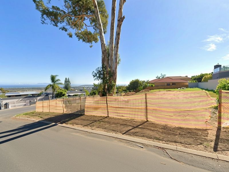 A vacant residential plot featuring a mature tree, bordered by a temporary construction fence, situated along a paved street with distant coastal views.