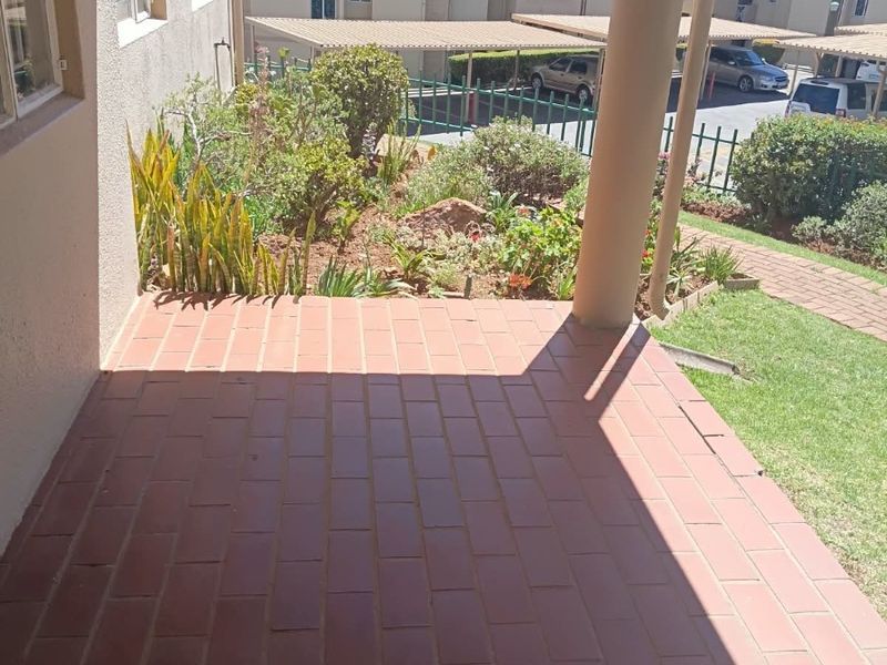A covered patio area featuring tiled flooring and a support column, overlooking a small garden and the townhouse complex parking area.